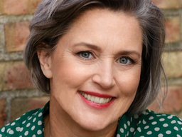 Margaret Price is smiling. They are standing in front of a brick wall and they are wearing a green top with white polka dots.