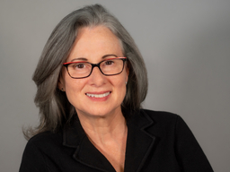 A woman with long gray hair and wearing eyeglasses and a black top is smiling.