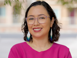 Yvette Martínez-Vu, a Chicana woman, is smiling. She is wearing a magenta-colored shirt and has dangly earrings.