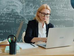 A white woman with blonde hair wearing a white shirt, black blazer, and black glasses sitting at a desk working on a computer. A chalkboard with equations written is in the background.