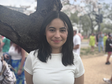 woman with dark, shoulder-length hair and white short sleeved t-shirt standing in front of tree.