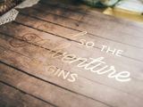 A wood table with the quote "and so the adventure begins"