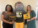 Two staff members posing with a cardboard cutout of the Retriever mascot. They hold signs saying I love UMBC and Retriever Nation.