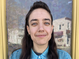 A white, non-binary, trans person with long dark hair, thick eyebrows, and a faint mustache smiling at the viewer. Their ears and nose are hooped with rings, and their watery button down shirt matches their light eyes. They are in front of a framed window that reflects a sunny Baltimore street.