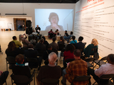 A gallery full of seated audience at an event where a speak stands at the front, a large video projection on the wall behind her