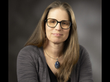 Professional photo of woman with long hair and glasses, large necklace, and gray sweater.
